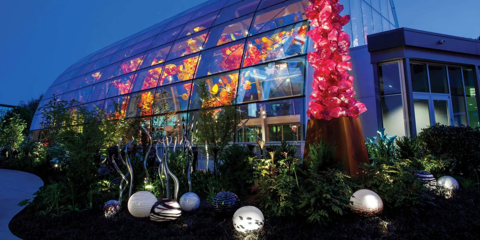 The Glasshouse seen from outside after dark, its curved glass wall filled with red and amber blown-glass forms